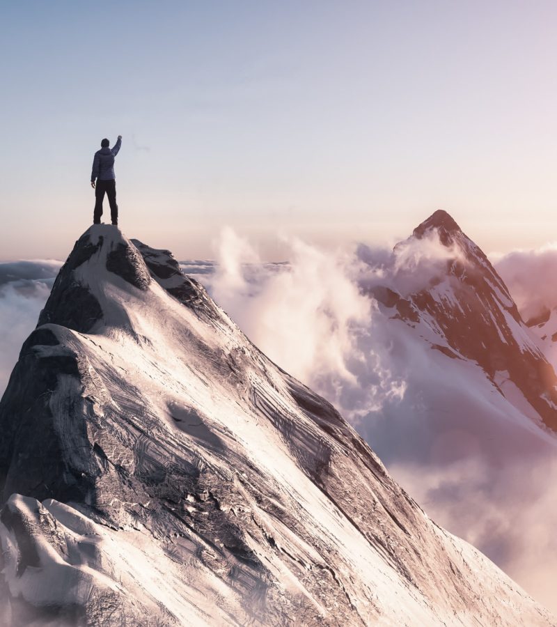 Adventure Composite. Adventurous Man is taking in the moment on top of a mountain. CGI Rocky Peak. Colorful Sunset or Sunrise Sky. Aerial Background Landscape from British Columbia, Canada.